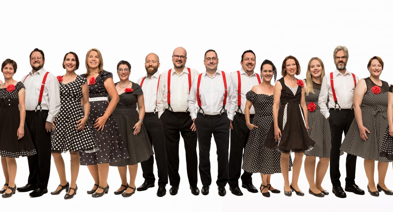 Ein gemischter Chor in loser Reihenfolge mit Damen in Rockabilly Kleidern, die Herren in schwarzen Hosen und weißen Hemden mit roten Hosenträgern