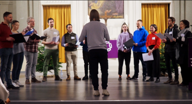 Eine Gruppe von Sängerinnen und Sängern steht im Halbkreis und singt gemeinsam Barbershop Harmonien.