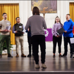 Eine Gruppe von Sängerinnen und Sängern steht im Halbkreis und singt gemeinsam Barbershop Harmonien.