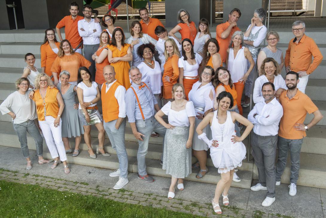 Eine Gruppe Menschen in orange, weiß, grau und silber gekleidet, steht auf einer Treppe und lacht in die Kamera.