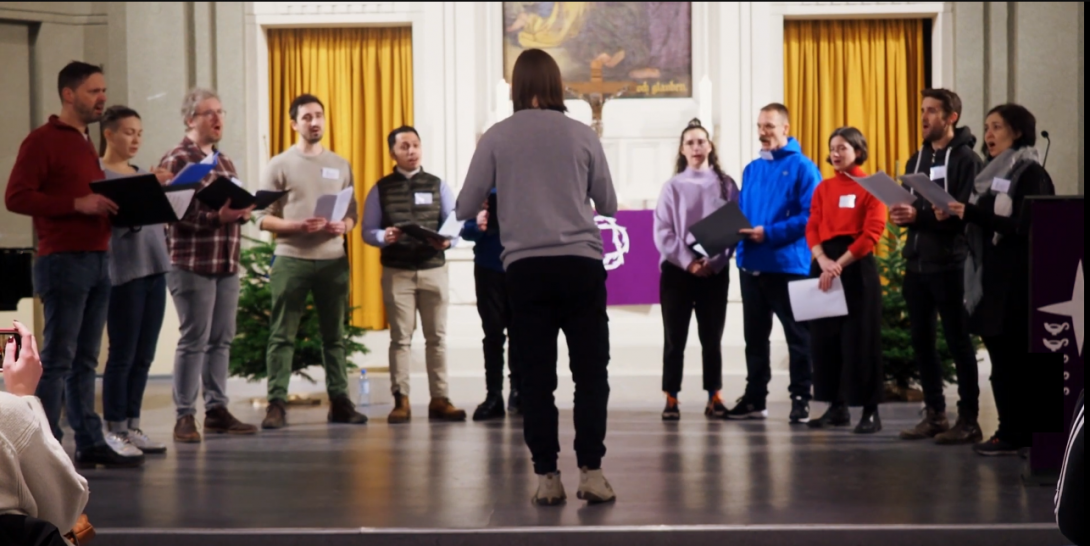 Eine Gruppe von Sängerinnen und Sängern steht im Halbkreis und singt gemeinsam Barbershop Harmonien.