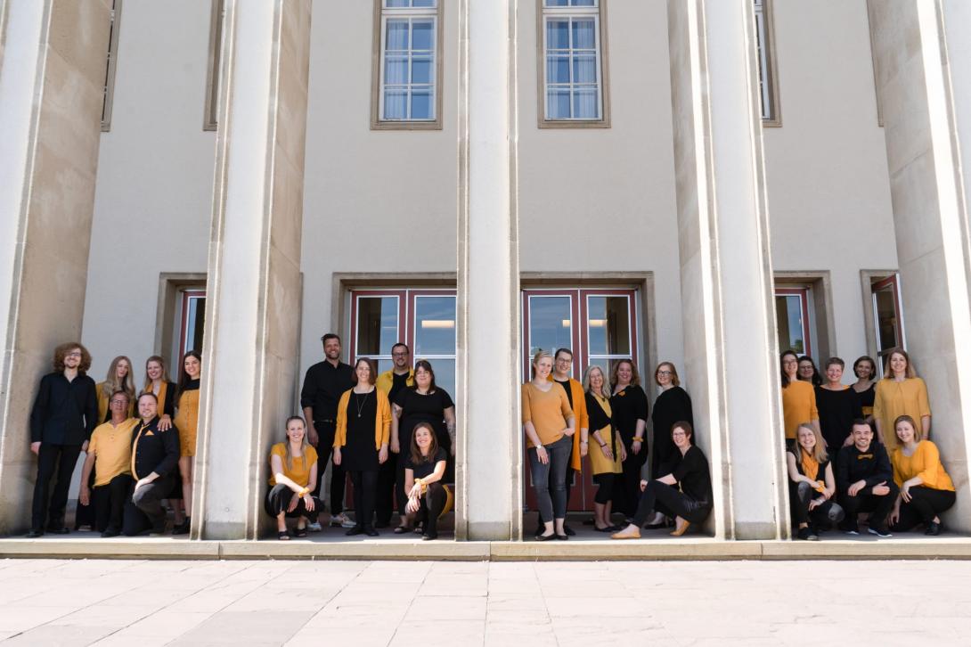 Ein Gruppenbild der Sänger:innen von B-Ware vor der Oetker-Halle in Bielefeld im Sommer 2024. Die Sänger:innen tragen ihre Chorfarben: Senfgelb und Schwarz.